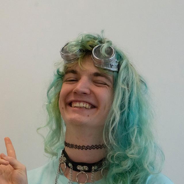 A person smiling widely with their eyes clothes. Their right hand is making a non-specific sign. They're wearing an aqua blue shirt, and they have blue-ish green hair that hits their shoulders. Above their eyes are some steampunk-style goggles. Their neck has a black choker with a black collar under it, which has multiple chains and rings on it